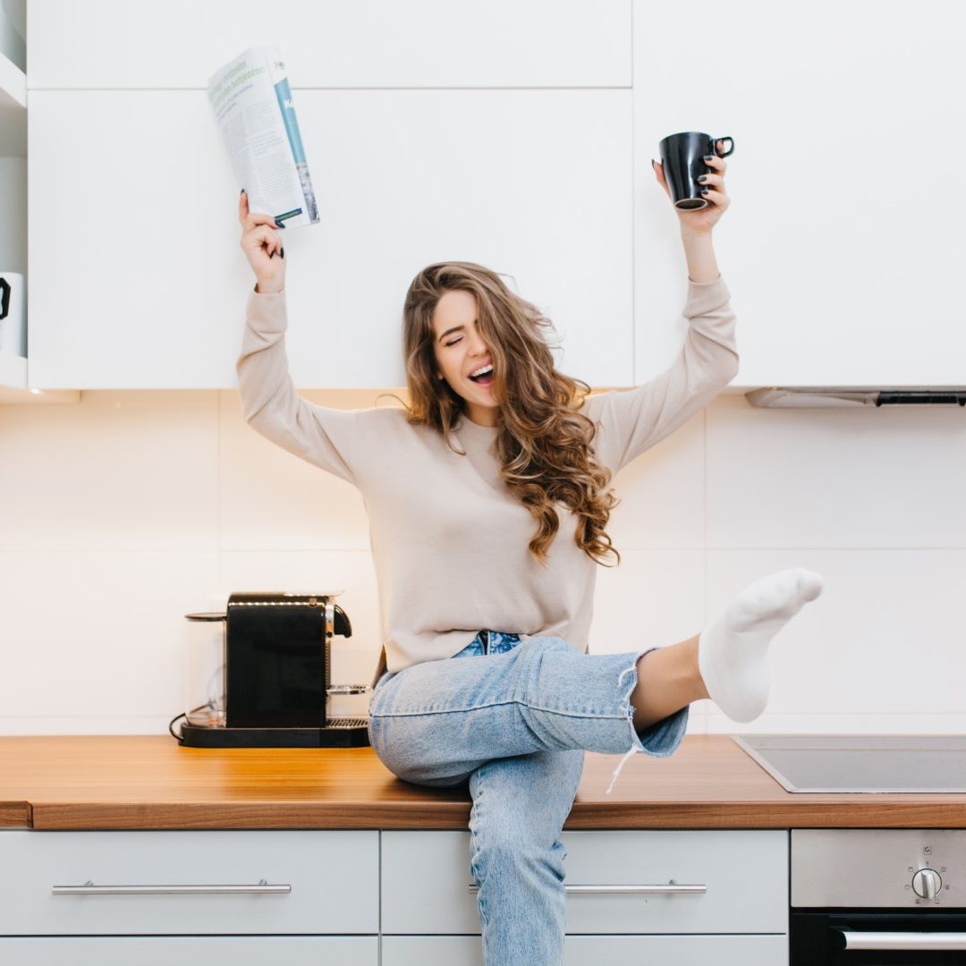 Graceful caucasian girl wears jeans enjoying good morning in her kitchen. Indoor portrait of emotional female model drinking coffee and reading magazine.